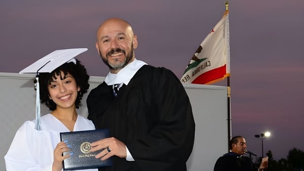 Un hombre y una mujer en toga de graduación están en una ceremonia de graduación en el Duarte Unified School District frente a una bandera.