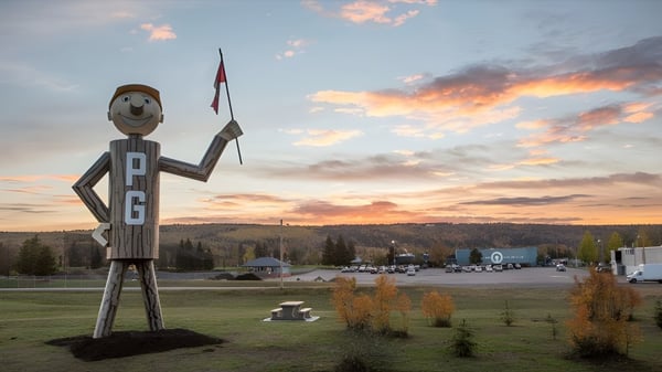 Una gran figura de madera con cara sonriente se encuentra frente a un paisaje con montañas y árboles en el terreno de la Duchess Park Secondary School.