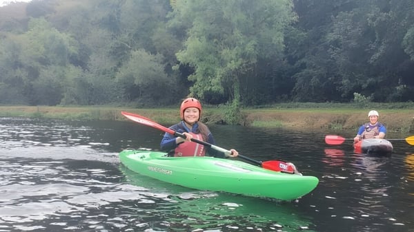 Dos alumnos del Duiske College reman en kayaks en un agua tranquila rodeada de árboles verdes.