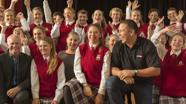 Un grupo de alumnas y alumnos en uniformes rojos está sentado y de pie juntos, al lado se sienta un hombre con camisa negra, en el campus de la Dunstan High School.