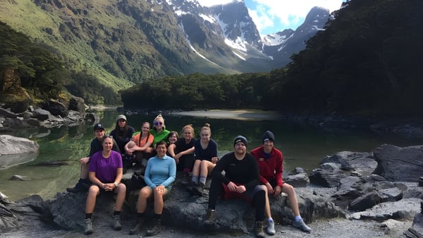 Un grupo de estudiantes está sentado en rocas en un paisaje montañoso con un lago y vegetación verde cerca de la Dunstan High School.