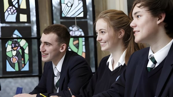 Tres estudiantes de la Durham School están de pie en uniforme escolar frente a una ventana de vitrales y sonríen.