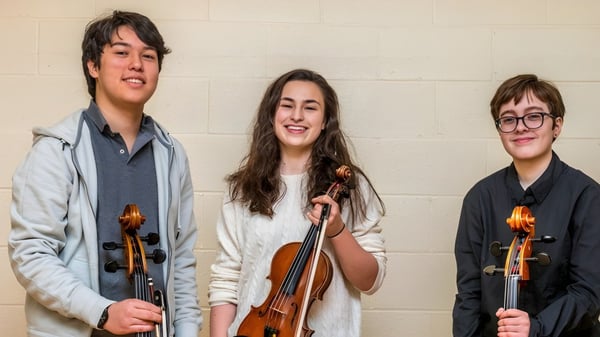 Tres estudiantes de la Eagle School sostienen instrumentos musicales y están frente a una pared sencilla.