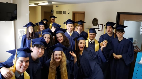 Un grupo de graduadas y graduados de la Eagle School en togas de graduación azules en una sala con televisor y muebles.