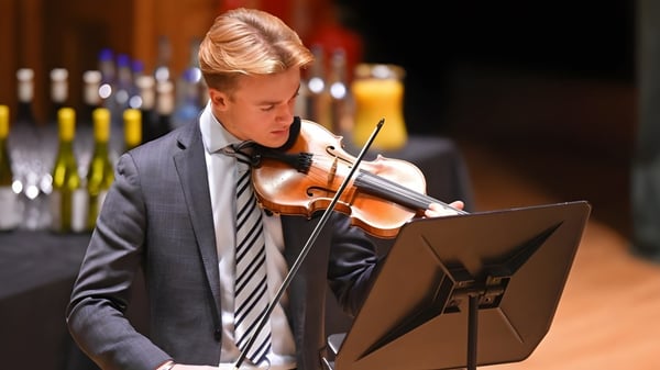Un joven vestido con un traje toca el violín en el escenario de la Earlscliffe School con botellas de vino de fondo.