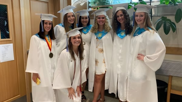 Un grupo de graduadas de la East Catholic High School en togas blancas con medallas se agrupan en el edificio escolar.