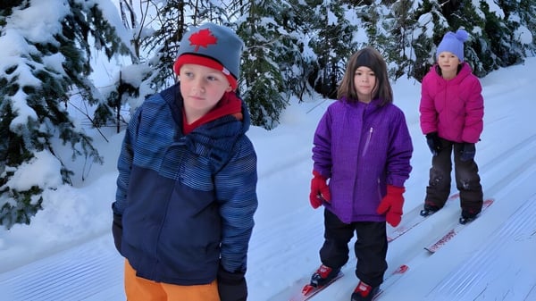 Tres personas con ropa de invierno cálida están en un camino nevado rodeadas de pinos cubiertos de nieve en el terreno de la École Franco-Nord.