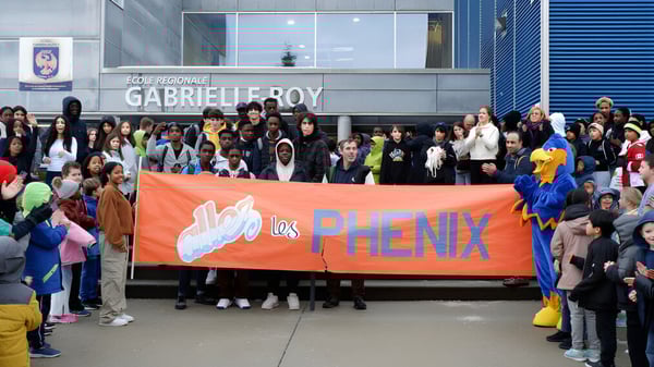 Estudiantes de la École Gabrielle-Roy sostienen un gran banner naranja con la inscripción Los Phenix frente al edificio escolar.