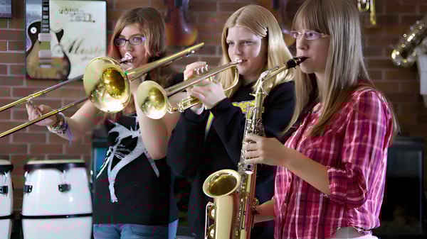 Tres estudiantes de la École J.E. Lapointe School tocan juntas instrumentos de metal en una sala con paredes de ladrillo.