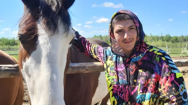 Una mujer está al lado de un gran caballo marrón en el área exterior de la École Marie-Esther frente a un fondo boscoso.