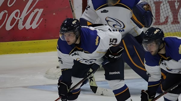Dos jugadores de hockey en uniforme juegan sobre el hielo frente a un anuncio de Coca-Cola en el terreno de la École Mathieu-Martin.