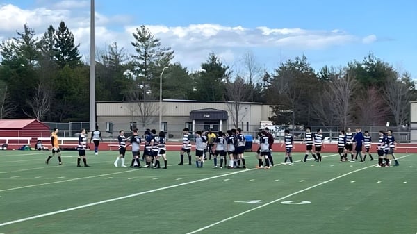 Un equipo deportivo de la École secondaire publique Mille-îles se reúne en un campo de césped con pinos y un edificio al fondo.