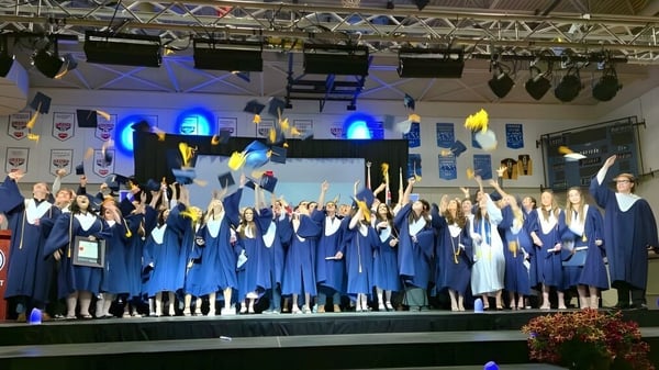 Los graduados de la École secondaire publique Le Sommet están en el escenario con sus togas de graduación azules y con iluminación de escenario de fondo.