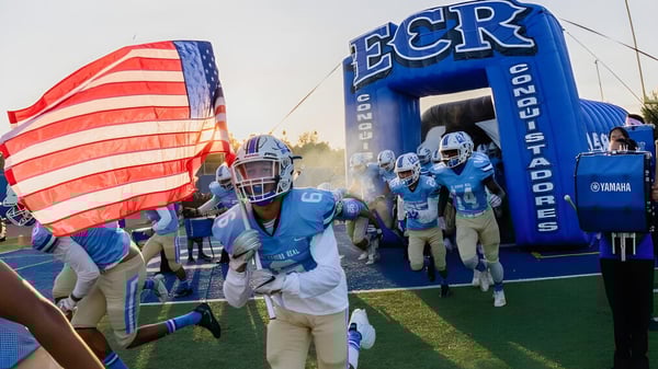 El equipo de fútbol de la El Camino Real Charter High School camina a través de un túnel inflable con la bandera americana en primer plano.