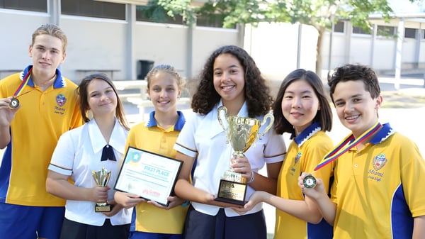 Un grupo de estudiantes de Elanora State High School sostiene trofeos y certificados frente al edificio escolar con árboles de fondo.