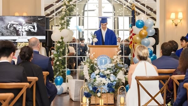 Un graduado da un discurso en la ceremonia de graduación de la Elite Preparatory Academy en un interior decorado festivamente.