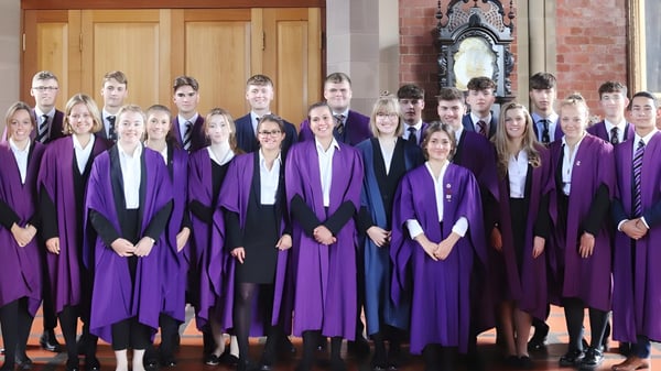 Un grupo de graduados está en túnicas moradas frente a un edificio de ladrillo con un arco decorado en el campus del Ellesmere College.