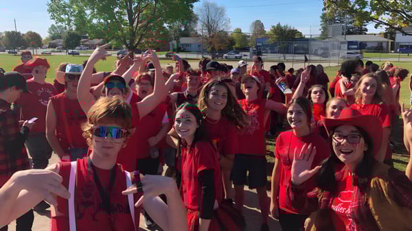 Un grupo de estudiantes de la Elmvale District High School lleva camisetas rojas y levanta los brazos con júbilo en un campo.