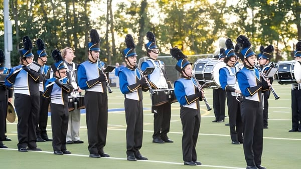 Miembros de la Marching Band de Elmwood High School están en formación en un campo de hierba frente a árboles y un cielo azul.