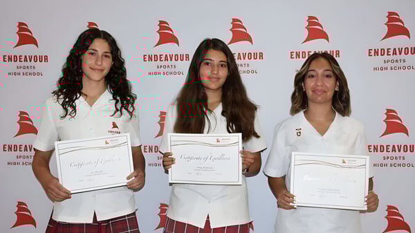 Tres alumnas de la Endeavour Sports High School sostienen sus certificados frente a un fondo con un logo.