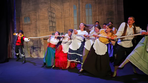 Un grupo de estudiantes del Epsom College lleva trajes tradicionales coloridos en un escenario con revestimiento de madera y ventanas de arco.