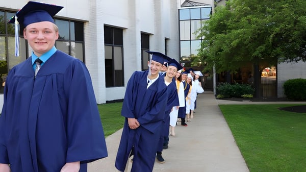 Un grupo de graduados está de pie en togas y birretes azules en el campus de la Erie First Christian Academy.