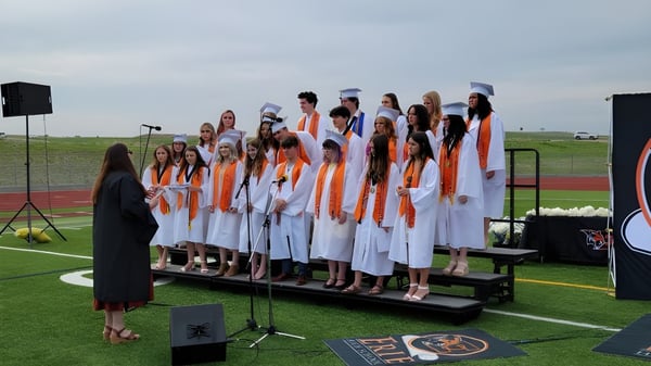 Un grupo de estudiantes de Erie High School está de pie en túnicas blancas y naranjas sobre un escenario al aire libre frente a altavoces.