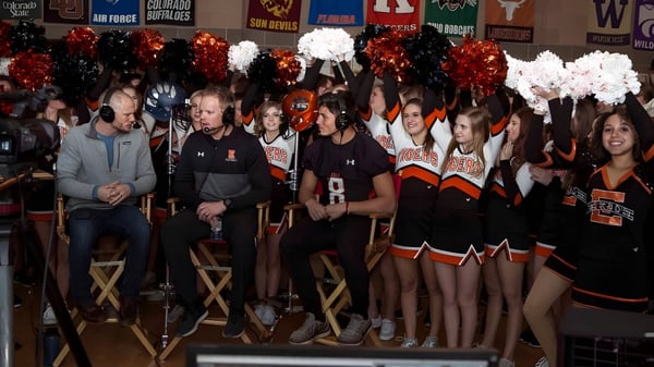 Un grupo de porristas en uniformes naranja y negro está de pie con pompones frente a los banners deportivos de Erie High School.