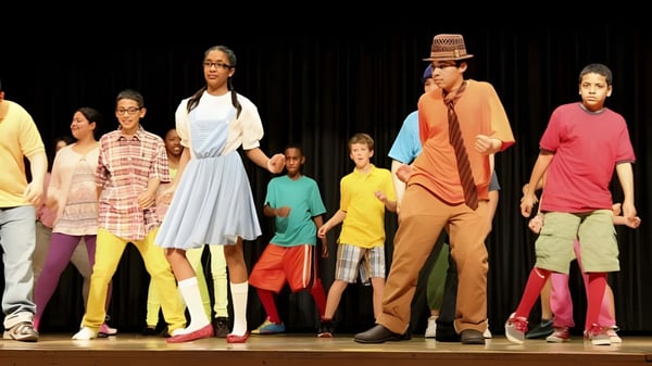Un grupo de estudiantes de la Erin District High School está en trajes coloridos sobre un escenario frente a un fondo oscuro.