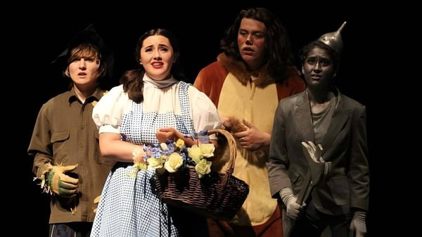 Un grupo de estudiantes de la Esquimalt High School está en trajes históricos con flores y cestas frente a un fondo oscuro.