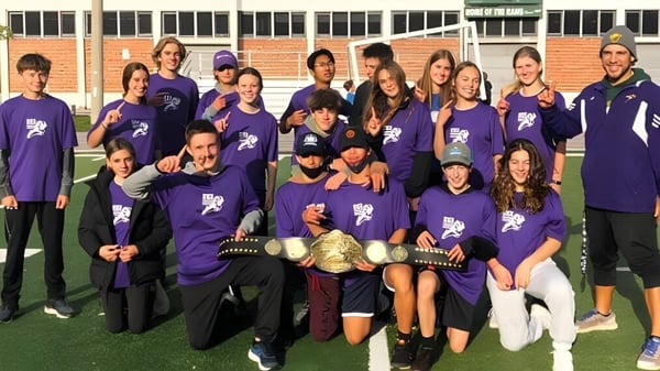 Un grupo de estudiantes en camisetas moradas está junto en el campo deportivo del Etobicoke Collegiate Institute frente a un edificio de ladrillo.