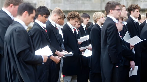 Un grupo de estudiantes del Eton College está de pie con ropa formal frente a un edificio de piedra sosteniendo documentos en la mano.