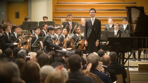 Una orquesta del Eton College se presenta en el escenario con un director y un pianista en el piano.