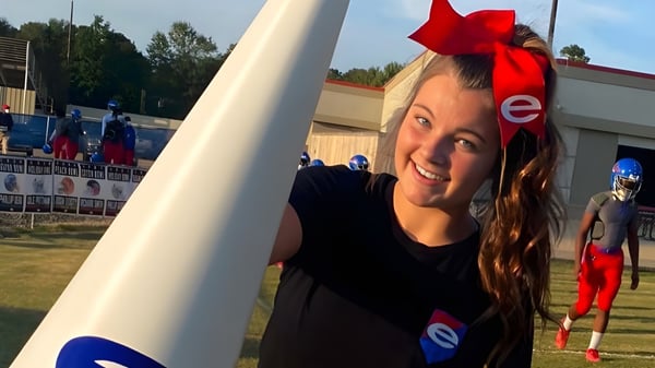Una joven con una cinta roja en el cabello sonríe frente al campo deportivo de la Evangel Christian Academy.