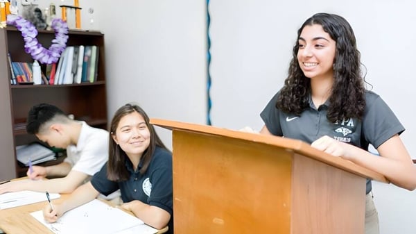 Tres estudiantes de la Fairmont Prep Academy están en el aula, una está en el podio, dos están sentadas en mesas.