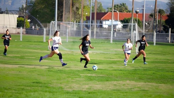 Jugadoras de fútbol de la Fairmont Prep Academy luchan en un campo con una portería frente a edificios y árboles.