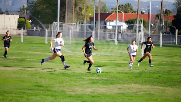 Un grupo de jugadoras de fútbol de la Fairmont Prep Academy juega en el campo de césped con una portería y edificios de fondo.