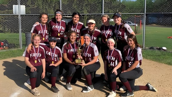 Un grupo de jóvenes jugadoras de softbol de la Faith Christian Academy (PA) posan en el campo de béisbol frente a una cerca de alambre.