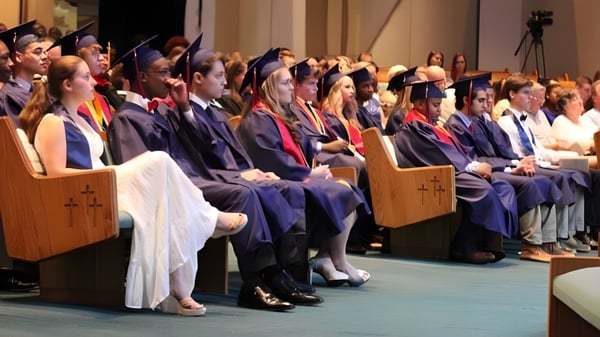 Graduados de la Faith Christian School están sentados en togas durante una ceremonia de graduación con escenario y público.