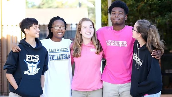 Un grupo de estudiantes de la Fayetteville Christian School está junto en camisetas deportivas al aire libre frente a una cerca con árboles.
