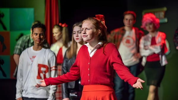 Un grupo de estudiantes actúa con vestidos rojos y blancos en el escenario frente a un fondo verde en el campus del Fingal Community College.