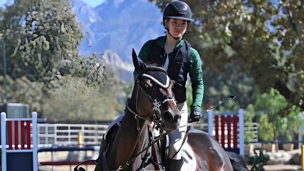 Una estudiante de la Flintridge Sacred Heart Academy monta un caballo al aire libre frente a árboles y montañas.