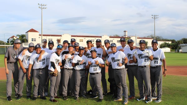 Un grupo de jugadoras y jugadores de béisbol de la Florida Preparatory Academy están en el campo de béisbol frente a un edificio con iluminación deportiva.