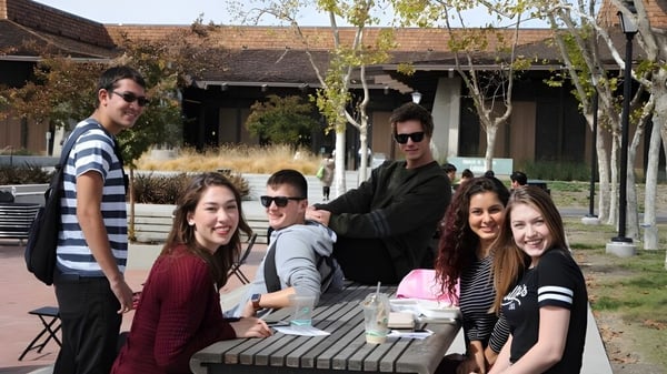 Un grupo de estudiantes se reúne en un deck de madera frente a un edificio rústico en el campus de Foothill College.