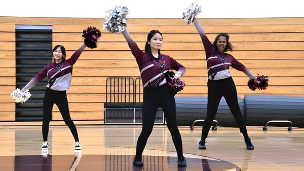 Tres estudiantes de la Fox Valley Lutheran High School están en la cancha de baloncesto en el gimnasio y practican una rutina de cheerleading con pompones.