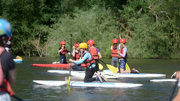 Estudiantes del Framlingham College están remando con cascos y chalecos salvavidas en tablas de paddle surf en un río.