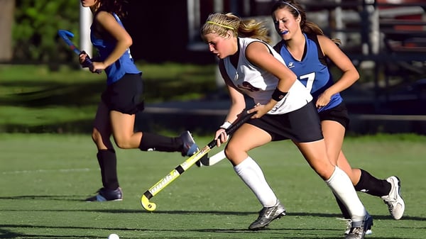 Estudiantes de la Frances Kelsey Secondary School juegan al hockey sobre césped en un campo deportivo con un estadio al fondo.