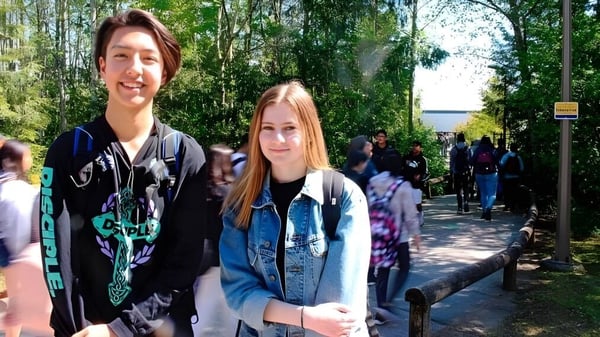 Dos estudiantes femeninas están afuera en el campus de la Fraser Heights Secondary School frente a árboles y una multitud.
