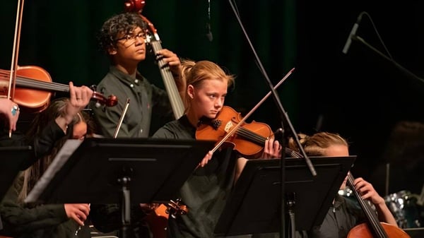 Un grupo de músicos de The Frederick Gunn School toca en el escenario; en primer plano se ve a un violinista y a un hombre con micrófono.