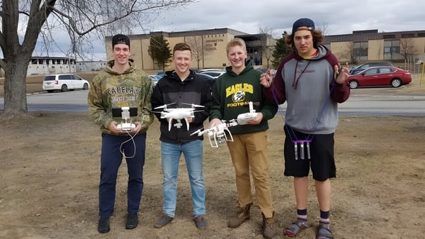 Un grupo de estudiantes se encuentra en un prado frente a los edificios del campus de la Fredericton High School.